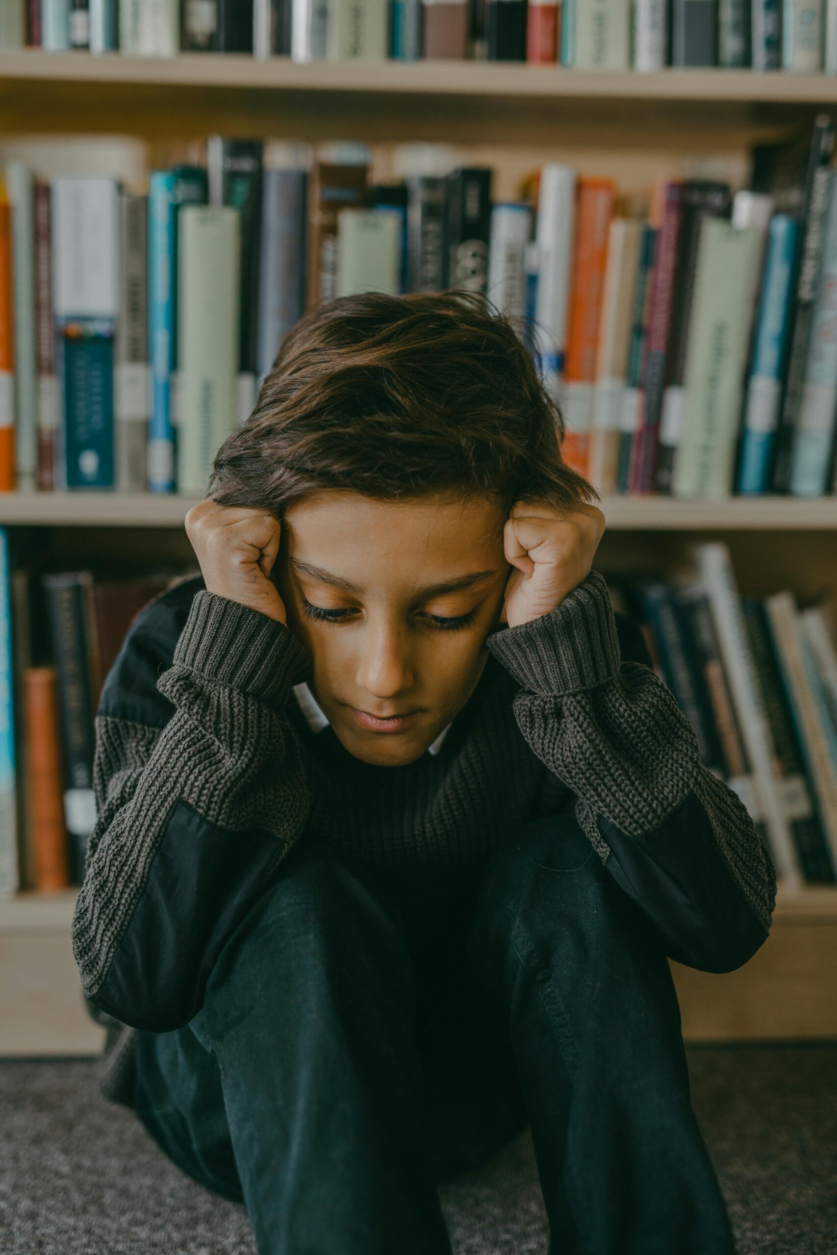 A distressed child sits on the floor in a library with books in the background, embodying sadness.